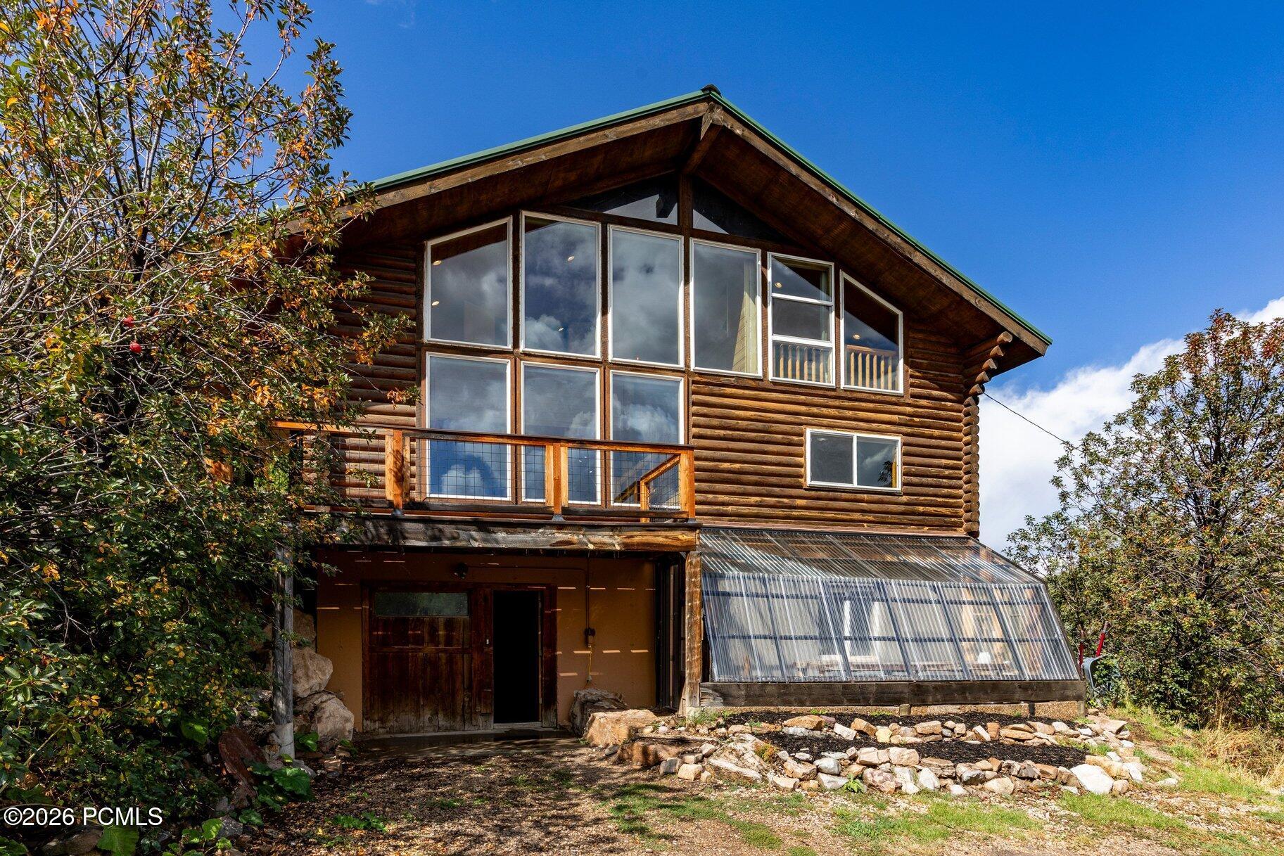 Log cabin with a peaked roof, large windows, balcony, and attached glass greenhouse.