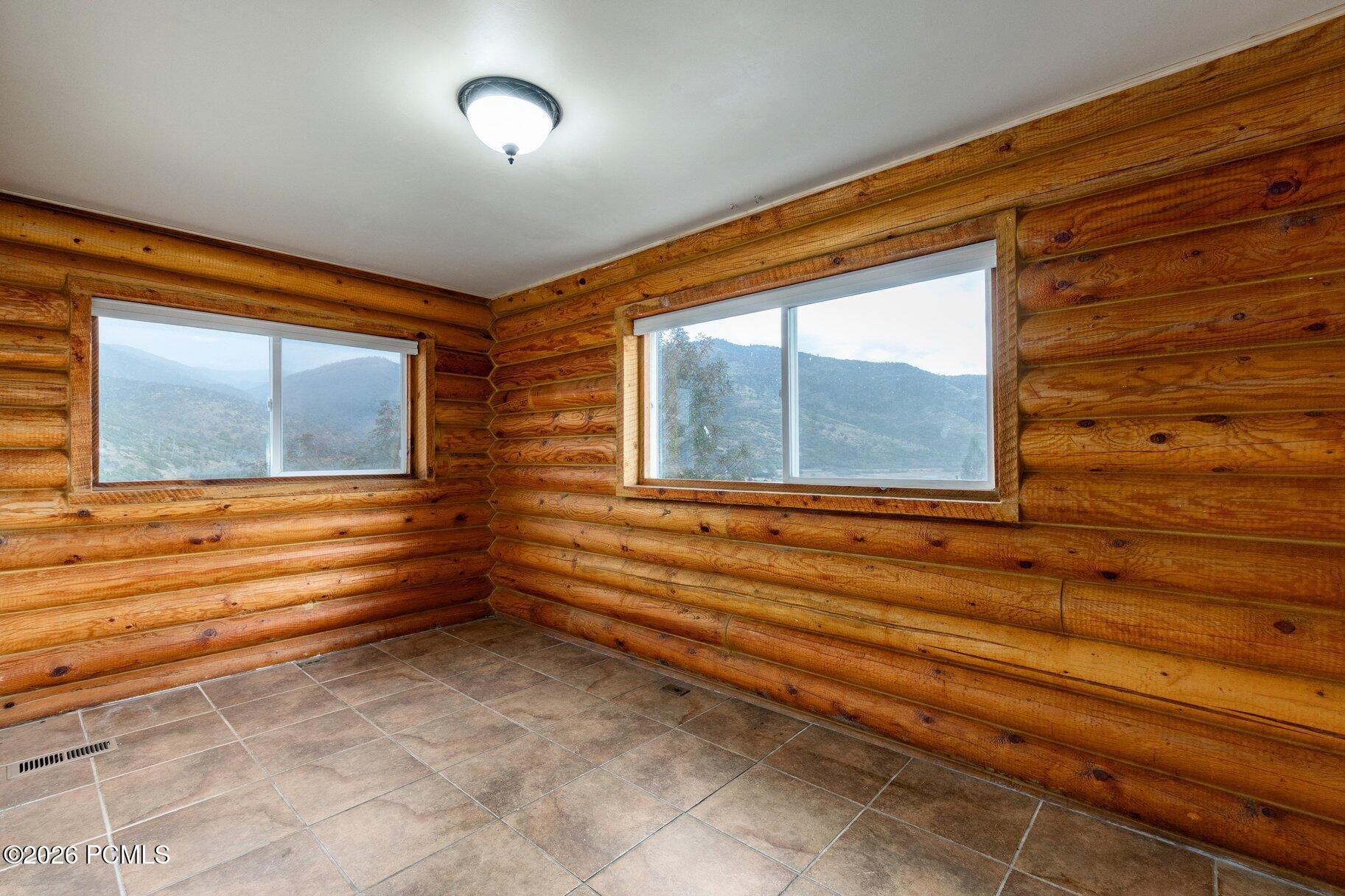 Empty log-walled room with tiled floor and mountain views through two windows.