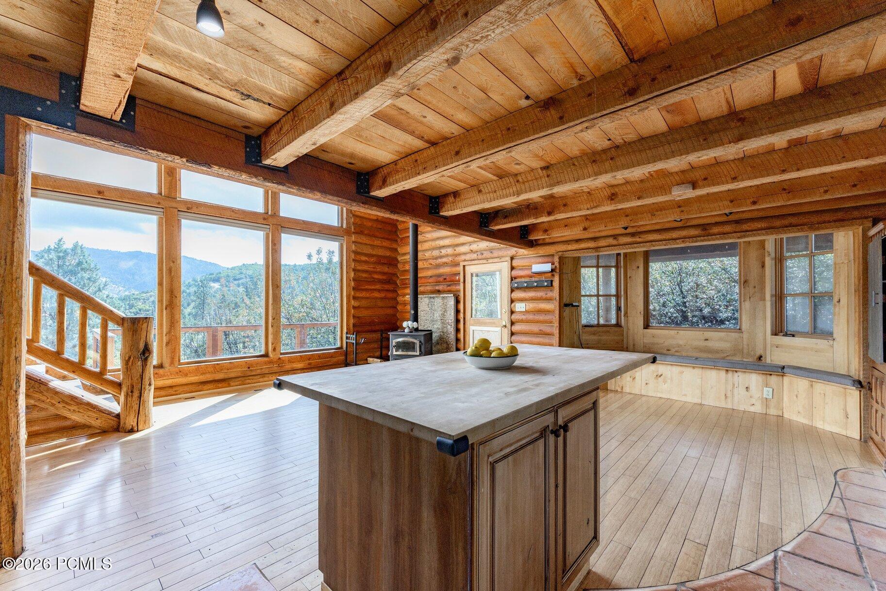 Rustic log-cabin kitchen with a large island, exposed beams, wood stove, and mountain views through broad windows.