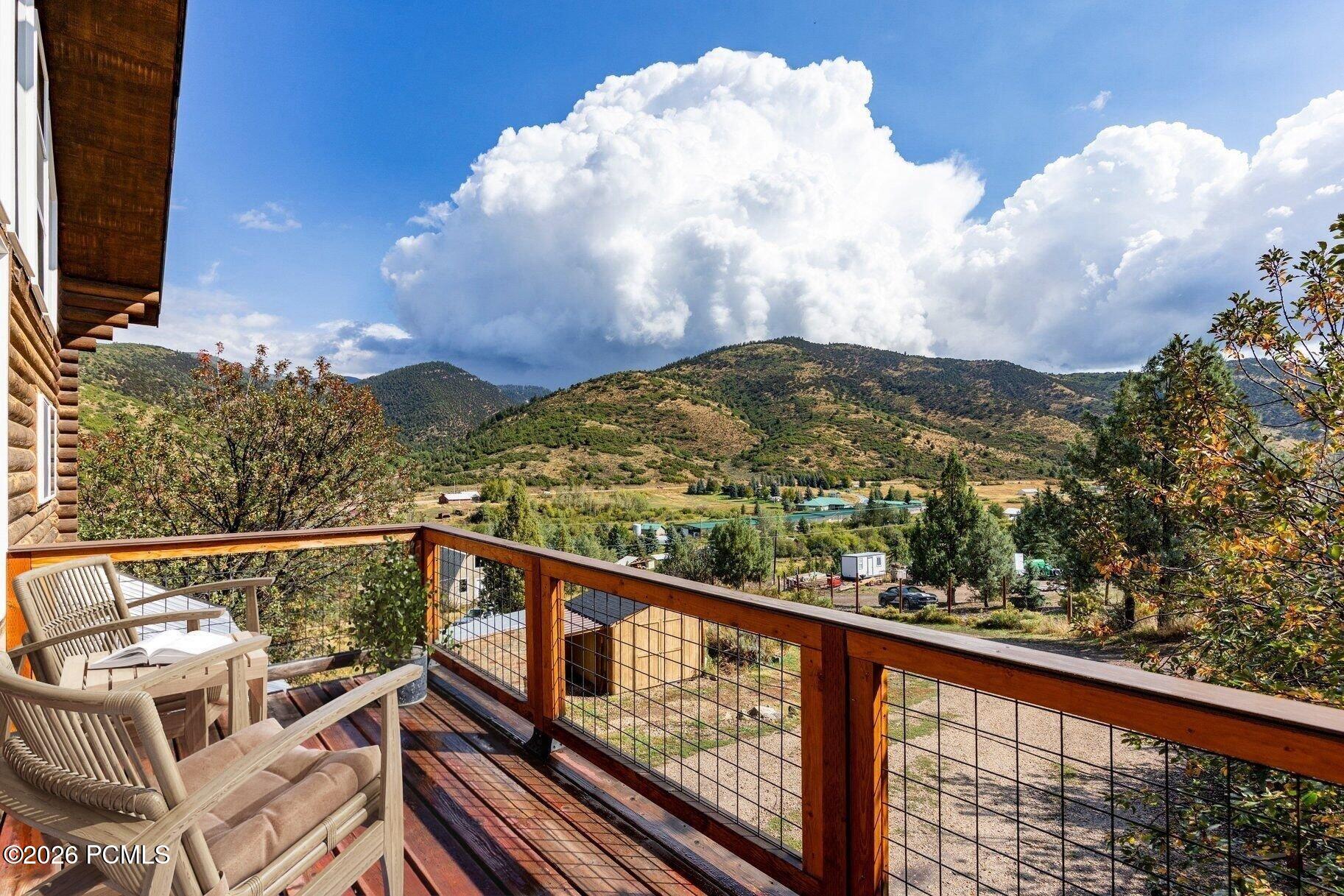 Wooden deck with Adirondack chairs overlooking mountain hills beneath a cloud-filled blue sky.