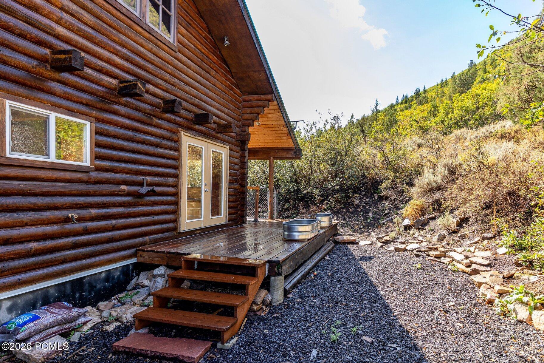 Log cabin exterior with a raised wooden deck and metal tubs beside a brush-covered hillside.