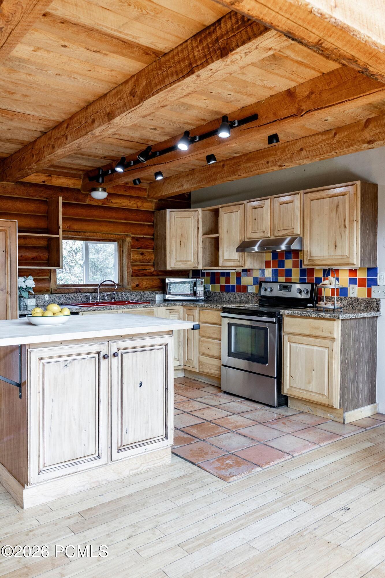 Rustic kitchen with exposed timber beams, log walls, pale wood cabinetry, and a colorful tile backsplash.