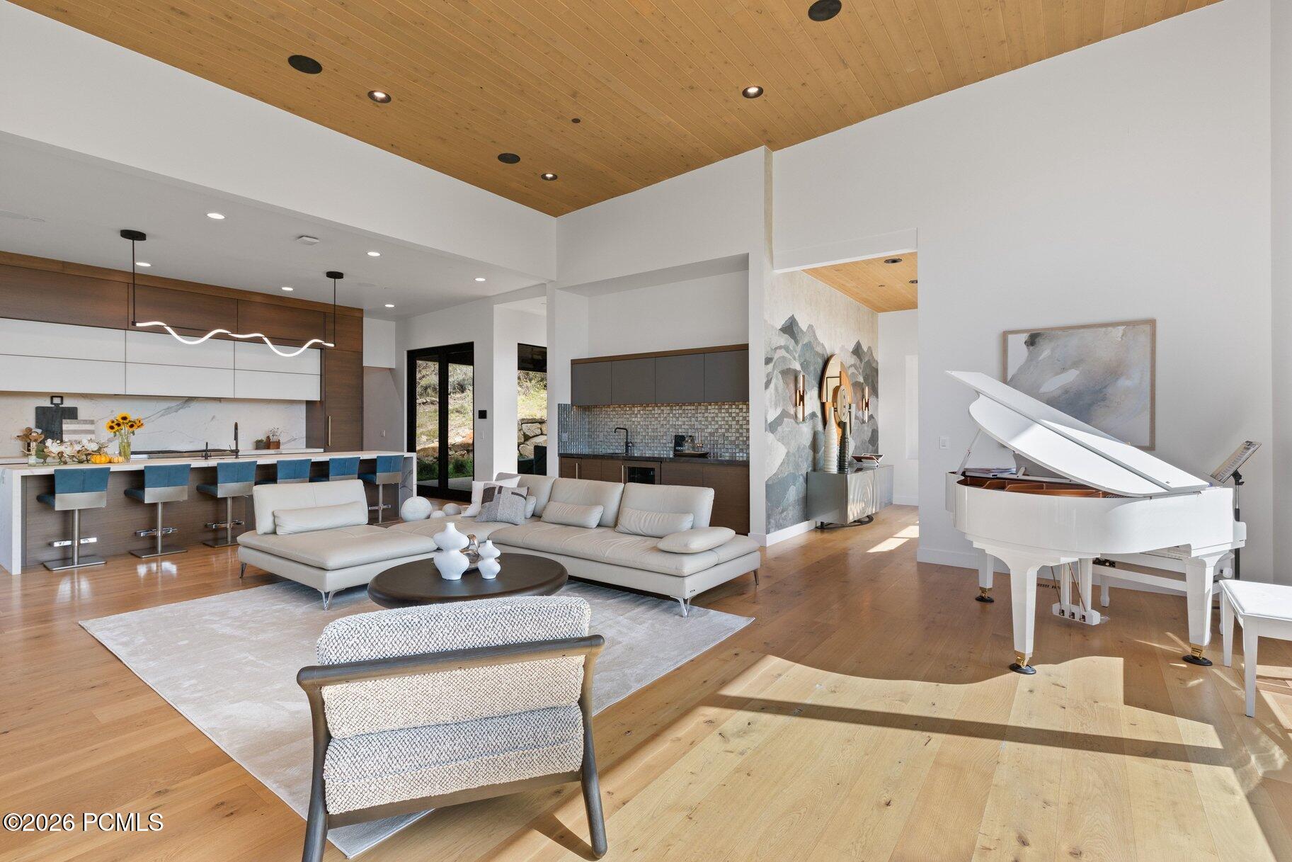Open-plan living room and kitchen with a white grand piano, sectional sofa, and wood-paneled ceiling.