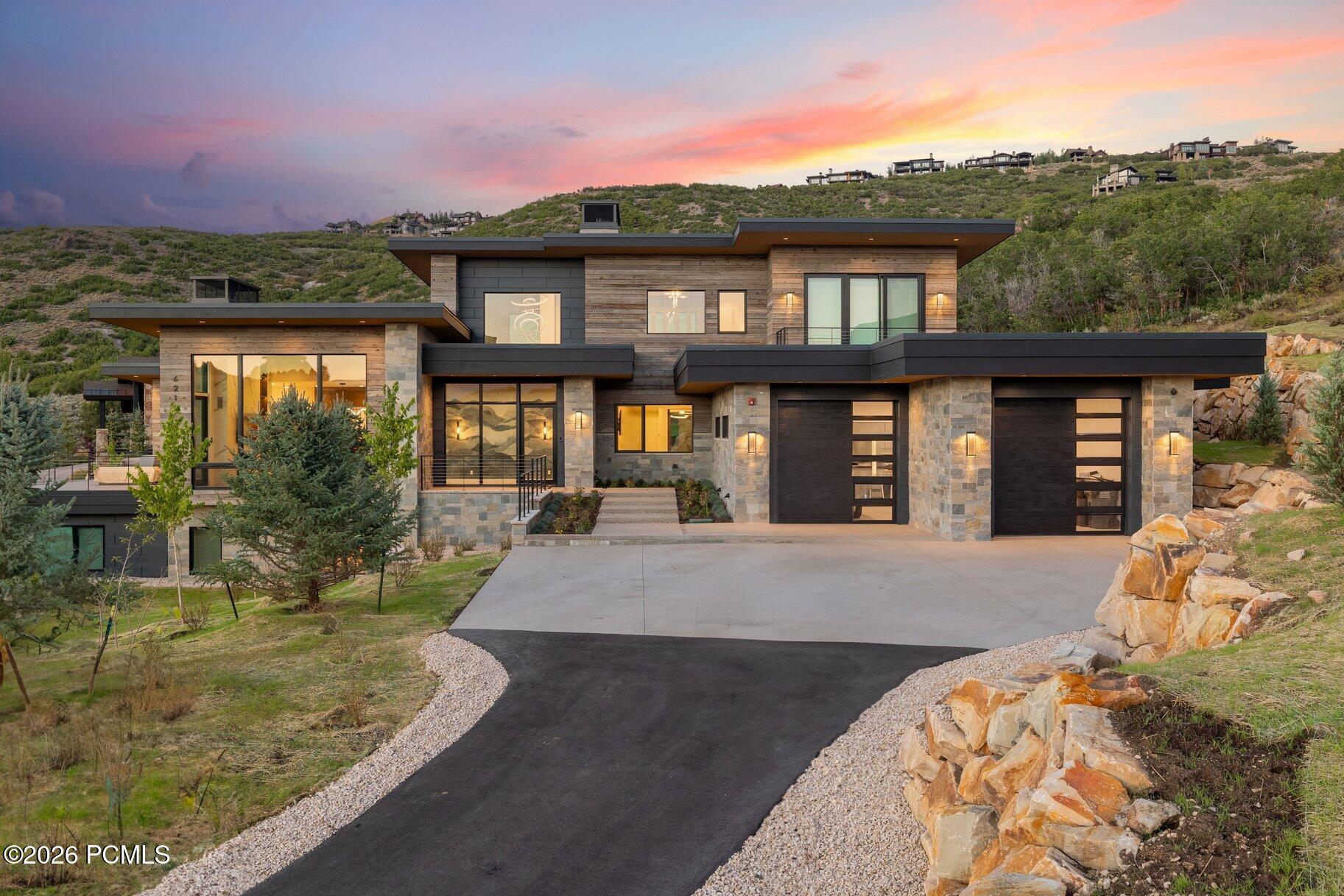 Modern wood-and-stone hillside home with a curving driveway beneath a pink dusk sky.