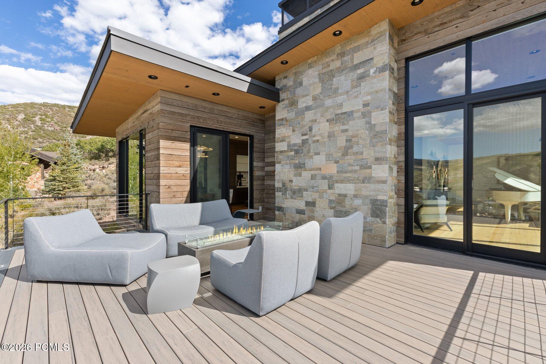 Furnished deck with gray lounge seating and a linear fire table beside a stone-clad modern home.