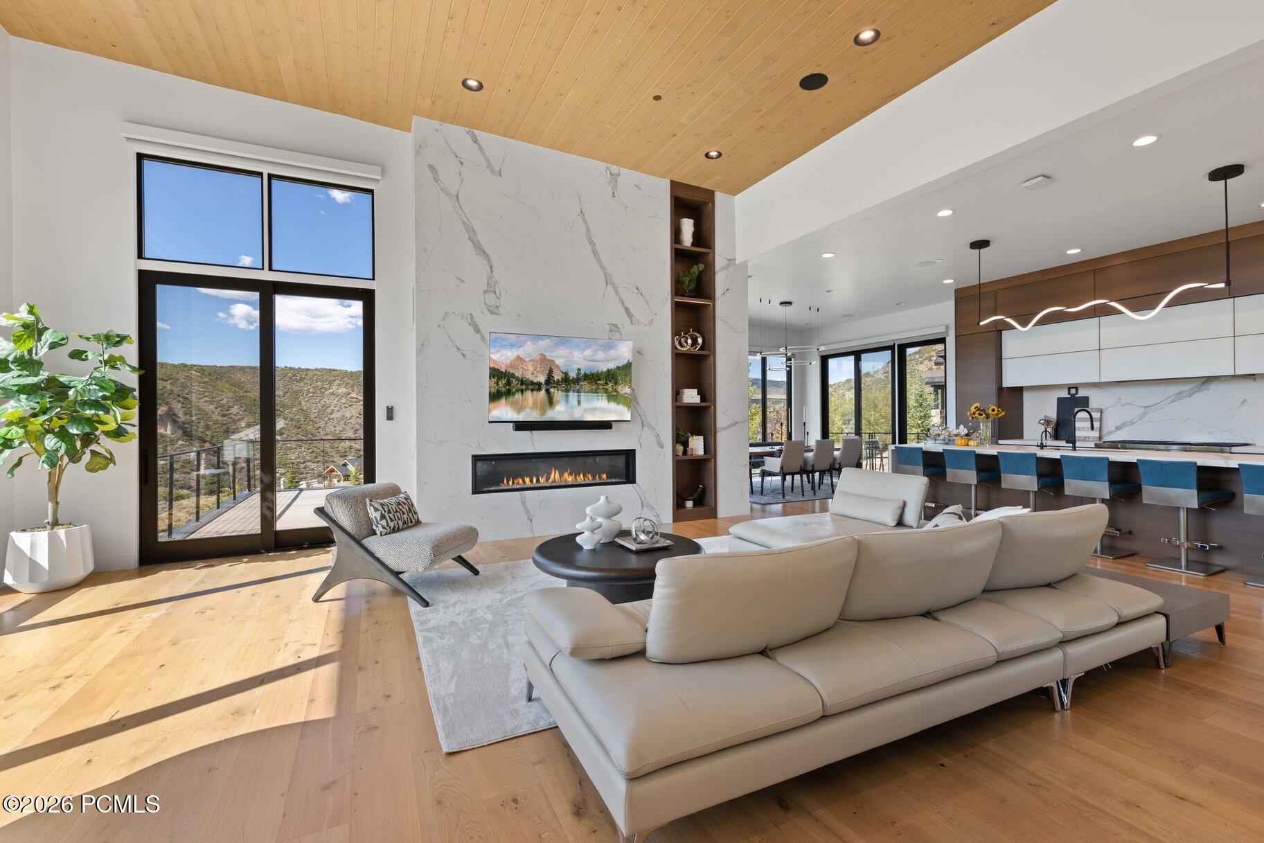 Modern living room with marble fireplace, wood ceilings, and large windows overlooking hills.