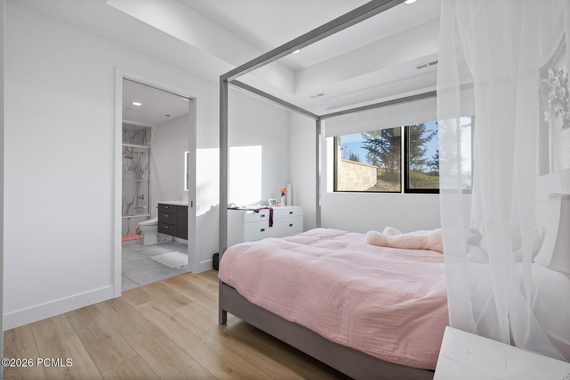 Bright bedroom with a pink-covered canopy bed, sheer curtains, and an ensuite bathroom doorway.