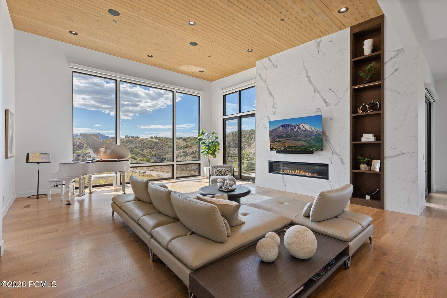 Modern living room with a white grand piano, marble fireplace, and expansive windows overlooking hills.