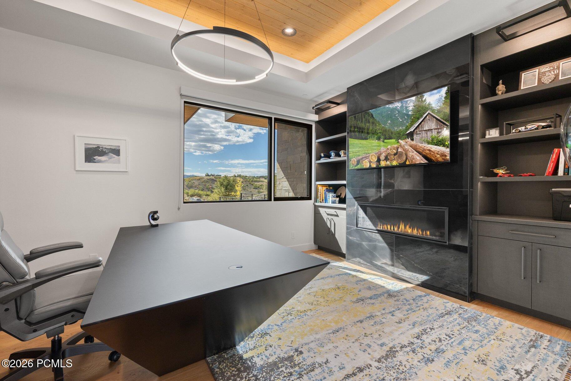 Furnished home office with a large desk, stone fireplace, wood ceiling, and hillside view.
