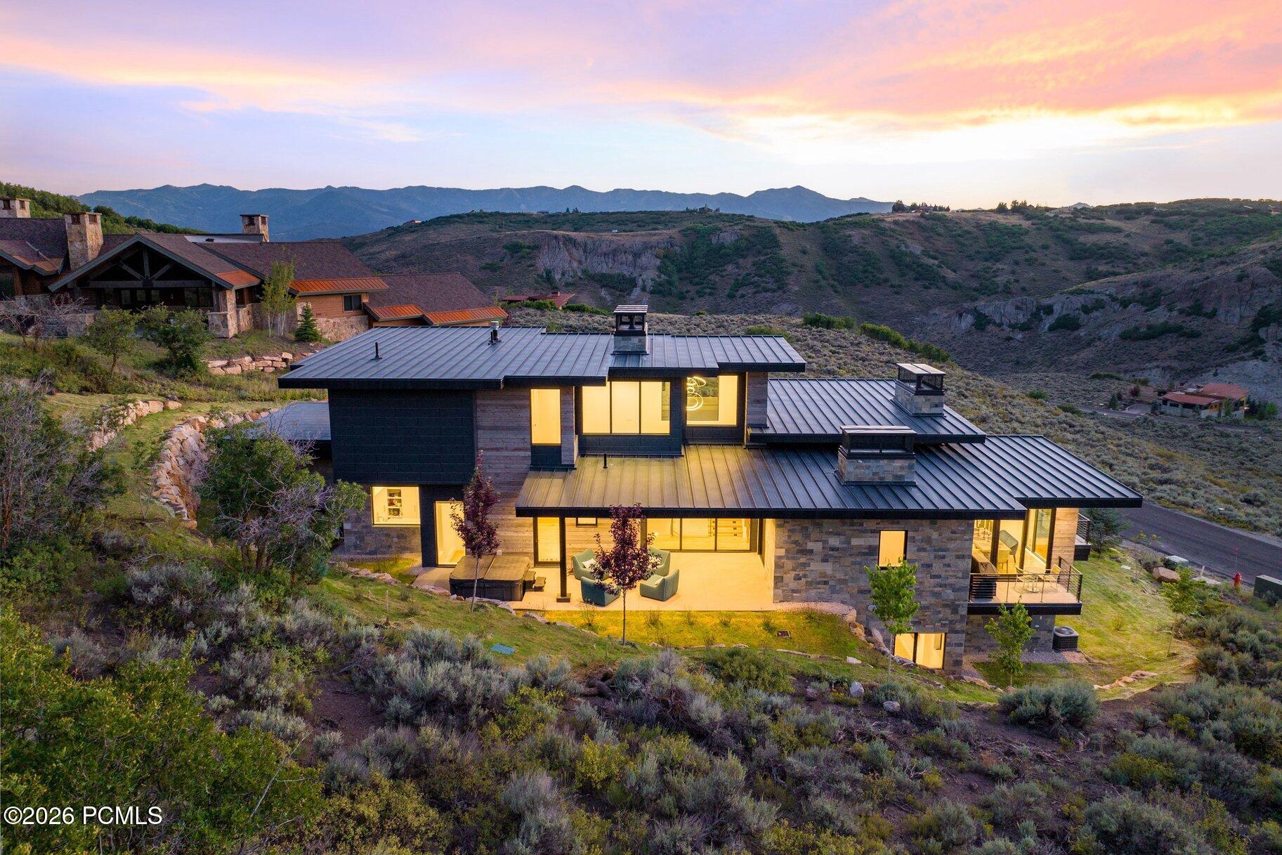 Modern hillside home with glowing windows and outdoor seating beneath a colorful sunset sky.