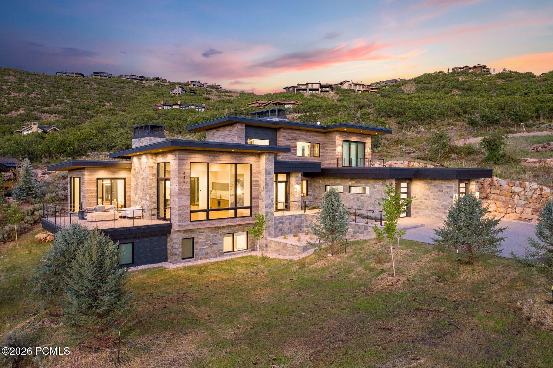Modern stone-and-wood hillside home with a furnished terrace beneath a colorful dusk sky.