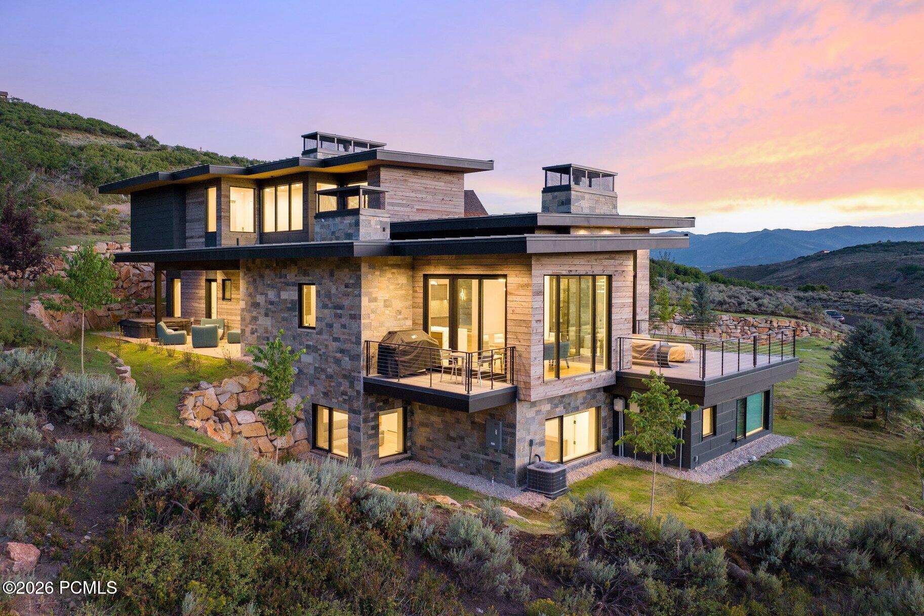 Modern stone-and-wood home with illuminated windows and terraces overlooking the mountains at sunset.