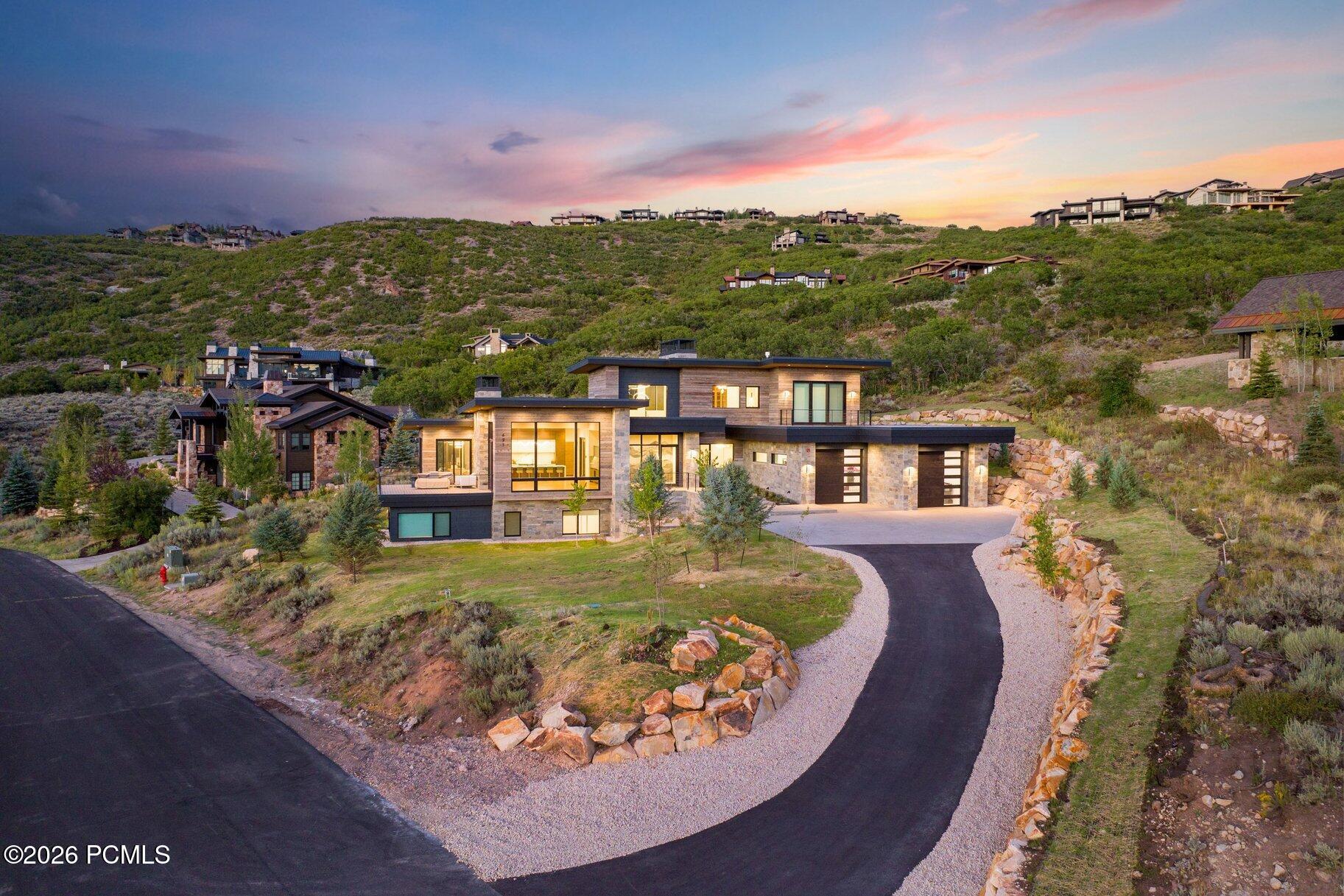 Modern hillside home with stone and wood exterior, curved driveway, and colorful sunset sky.