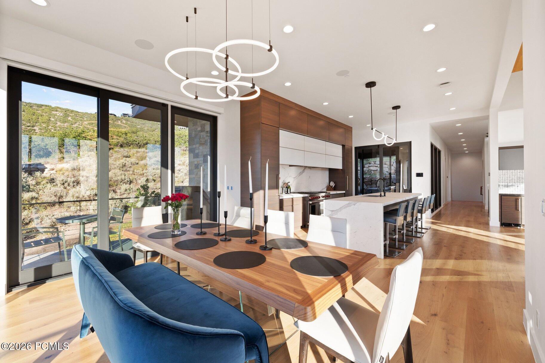 Open-plan dining room and kitchen with a wood table, marble island, and hillside views through glass doors.