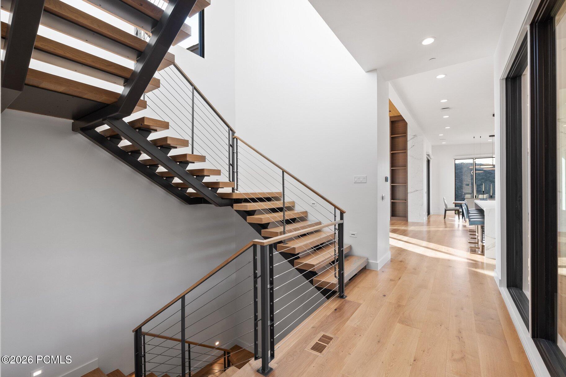 Modern home staircase with wood treads and dark metal framing beside a bright wood-floored hallway.