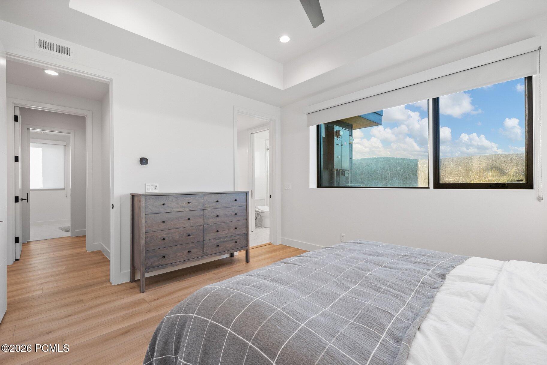 Bright bedroom with a gray quilted bed, wood dresser, and a large window overlooking wooded hills.