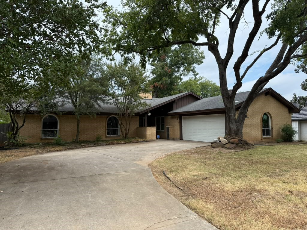 Single-story brick home with arched windows, attached garage, wide driveway, and mature shade trees.