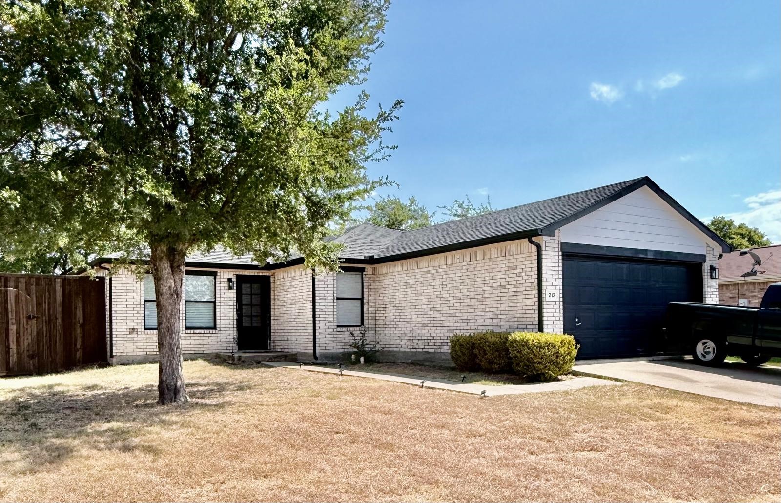 Single-story brick house with a dark garage door, large shade tree, and dry front lawn.