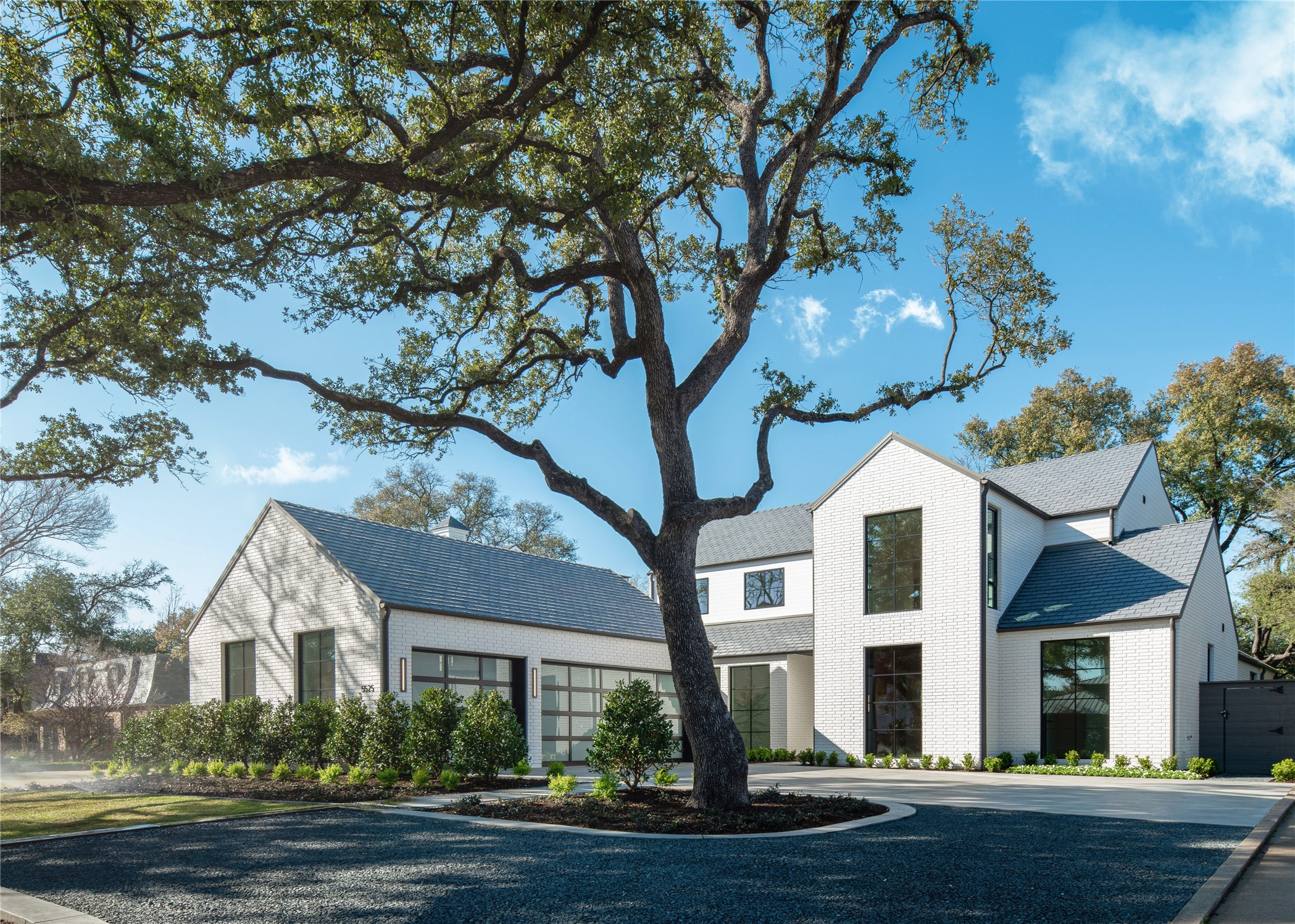 Modern white-brick home beneath a large mature oak, with dark-framed windows and gravel drive.