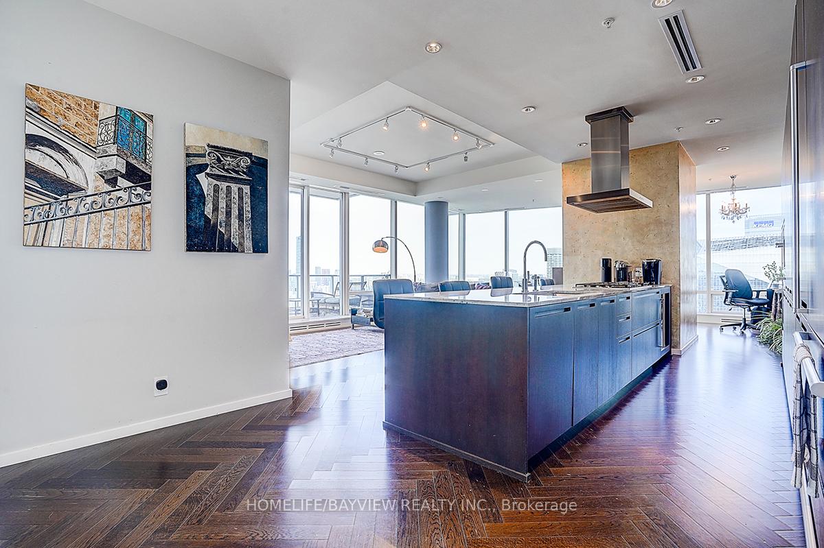 Open-plan penthouse kitchen with a large blue island, herringbone floors, and floor-to-ceiling city-view windows.