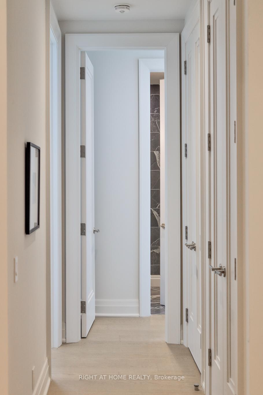 Bright hallway with pale wood floors, white paneled doors, and a glimpse of a marble-tiled bathroom.
