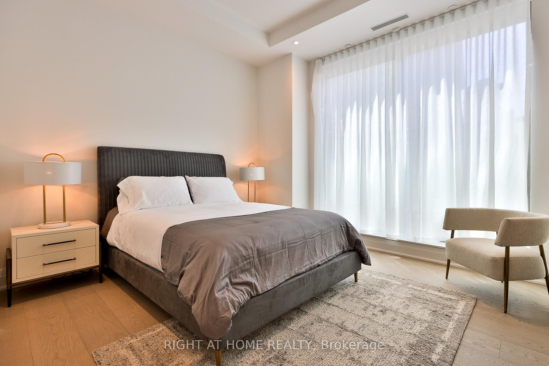 Staged bedroom with a gray upholstered bed, sheer curtains, and an armchair beside a large window.