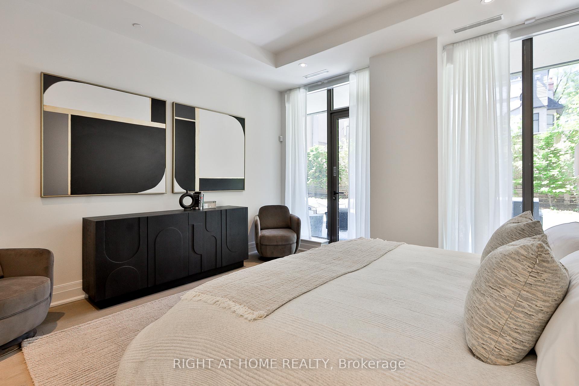 Bedroom with neutral bedding, abstract wall art, and tall windows overlooking a garden patio.