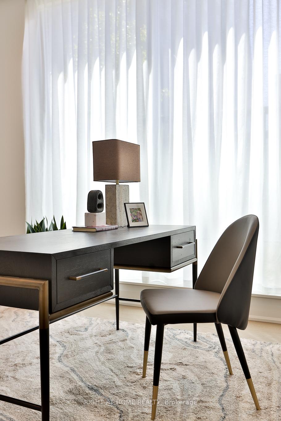 Furnished home office with a dark desk and chair in front of tall sheer-curtained windows.