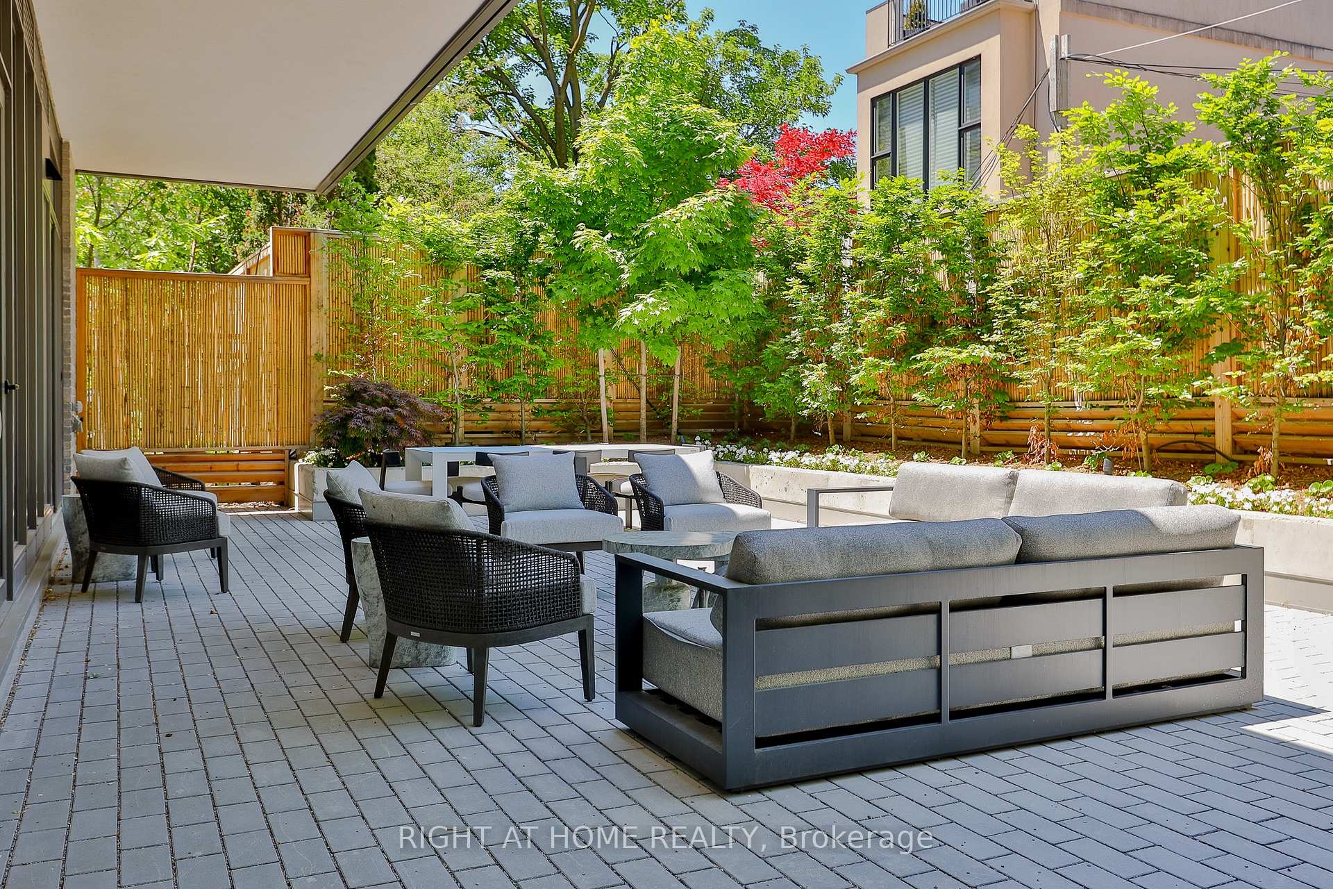 Furnished garden patio with modern seating, gray pavers, bamboo fencing, and leafy trees.