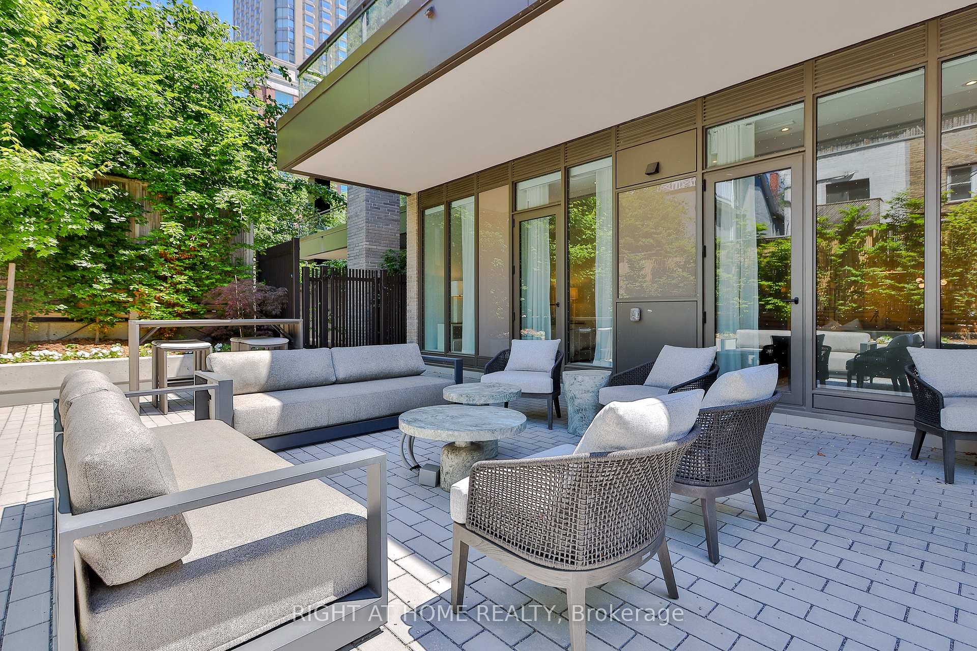 Furnished garden terrace with gray sofas, woven chairs, and leafy trees beside a modern building.