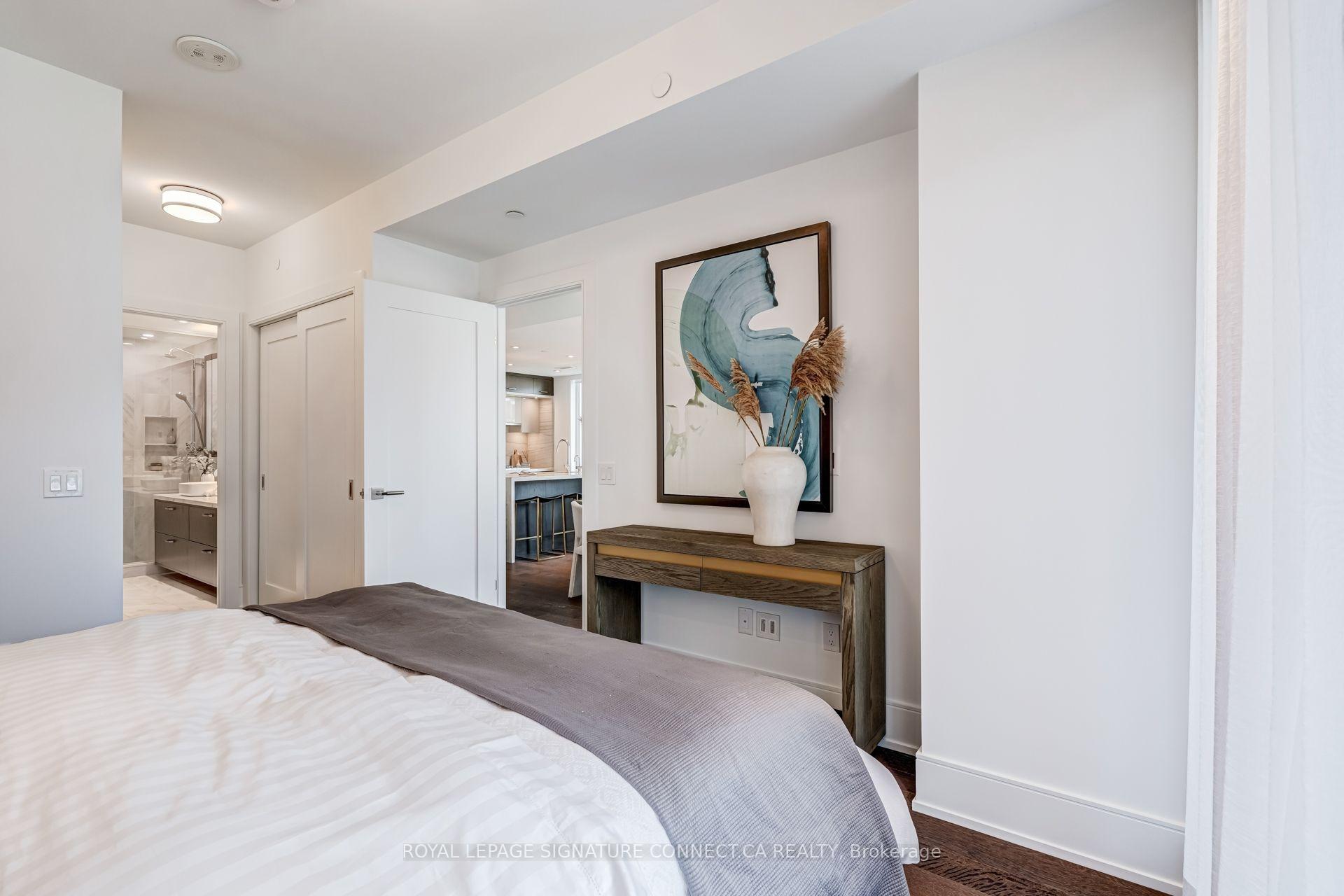 Staged bedroom with a large bed, wood console, abstract blue artwork, and glimpses of the ensuite and kitchen.
