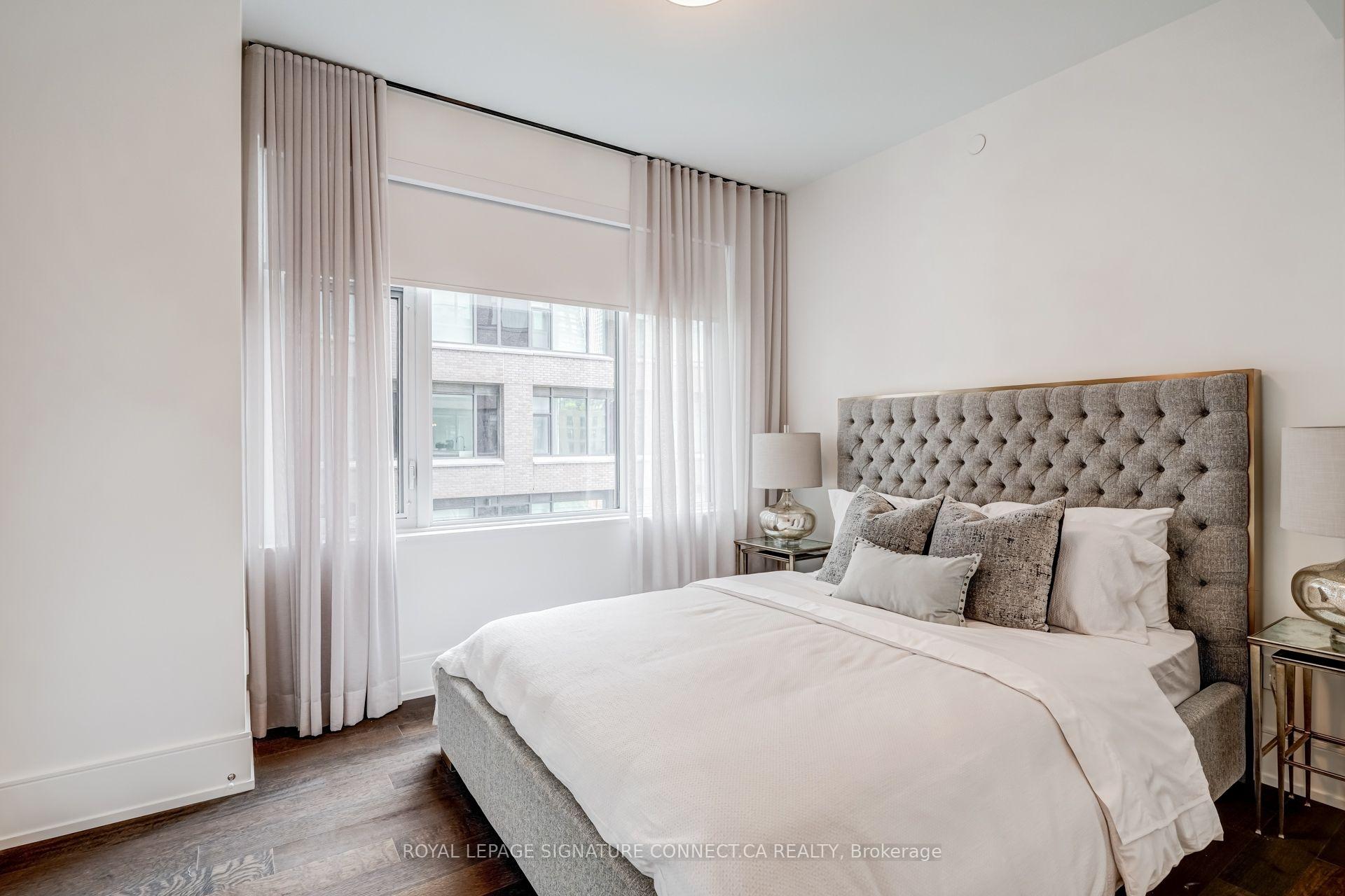Staged condo bedroom with a gray tufted headboard, white bedding, and tall curtains beside a large window.