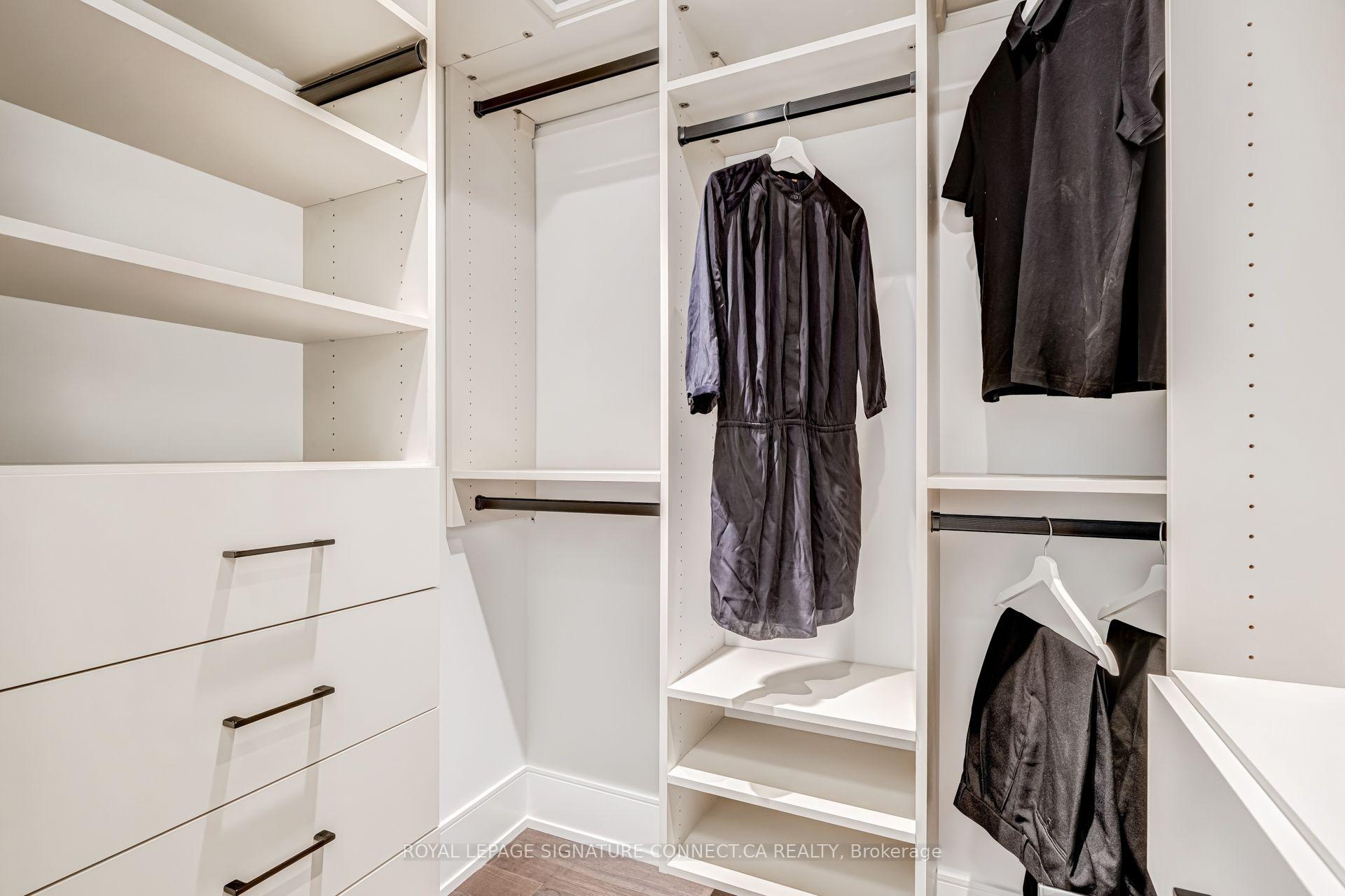 Walk-in closet with white built-in shelving and drawers, hanging rails, and dark clothing.
