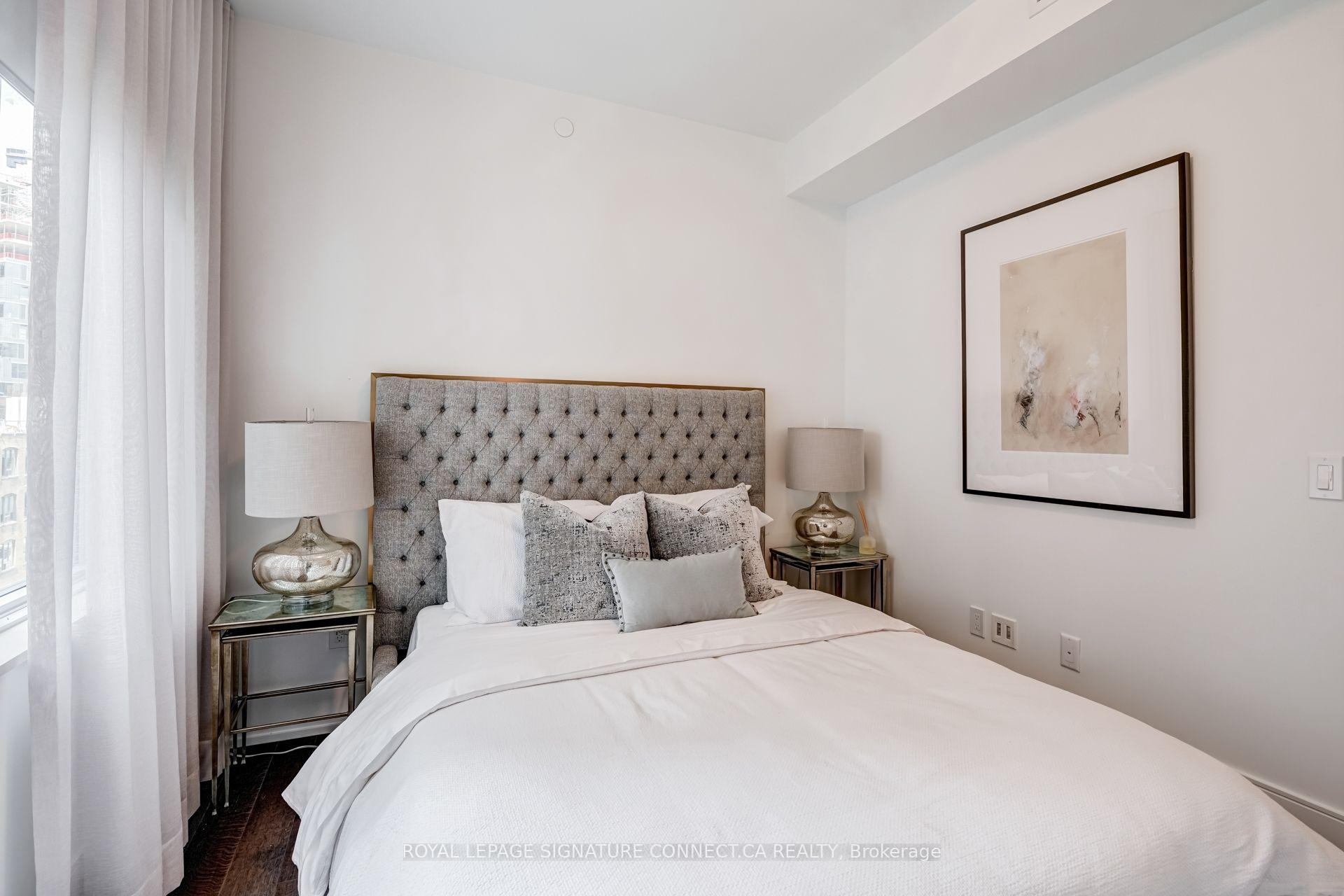 Staged bedroom with a white bed, gray tufted headboard, sheer window curtains, and framed abstract art.