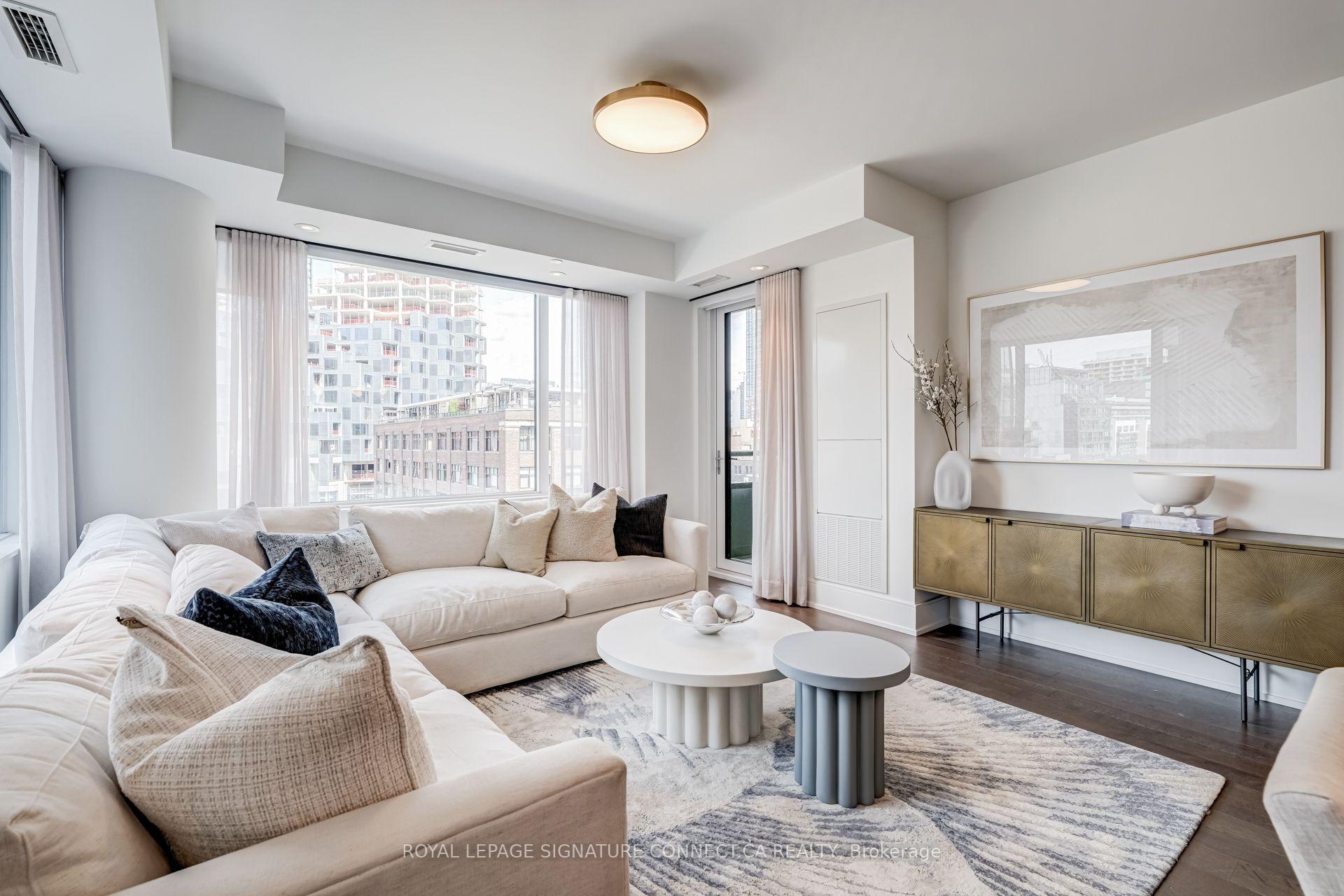 Staged condo living room with a cream sectional, round coffee tables, and large windows overlooking city buildings.