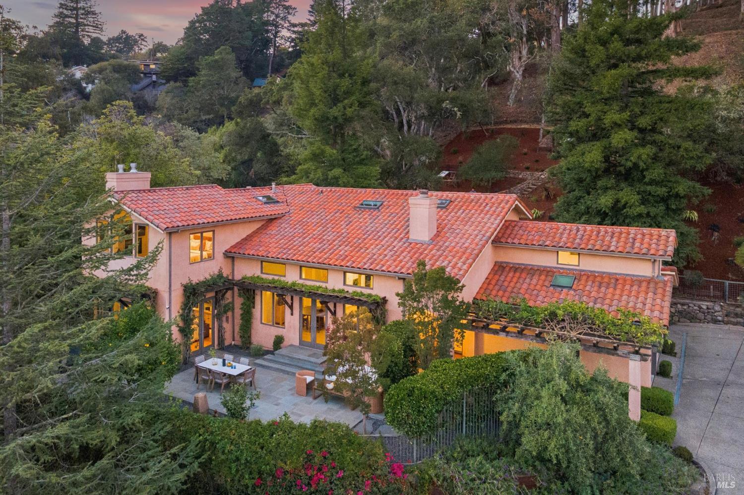 Pink-stucco hillside home with a terracotta tile roof, landscaped patio, and warmly lit windows at dusk.