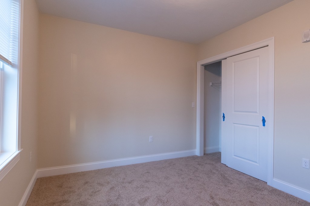 Empty bedroom with beige carpet, a window with blinds, and an open sliding closet.