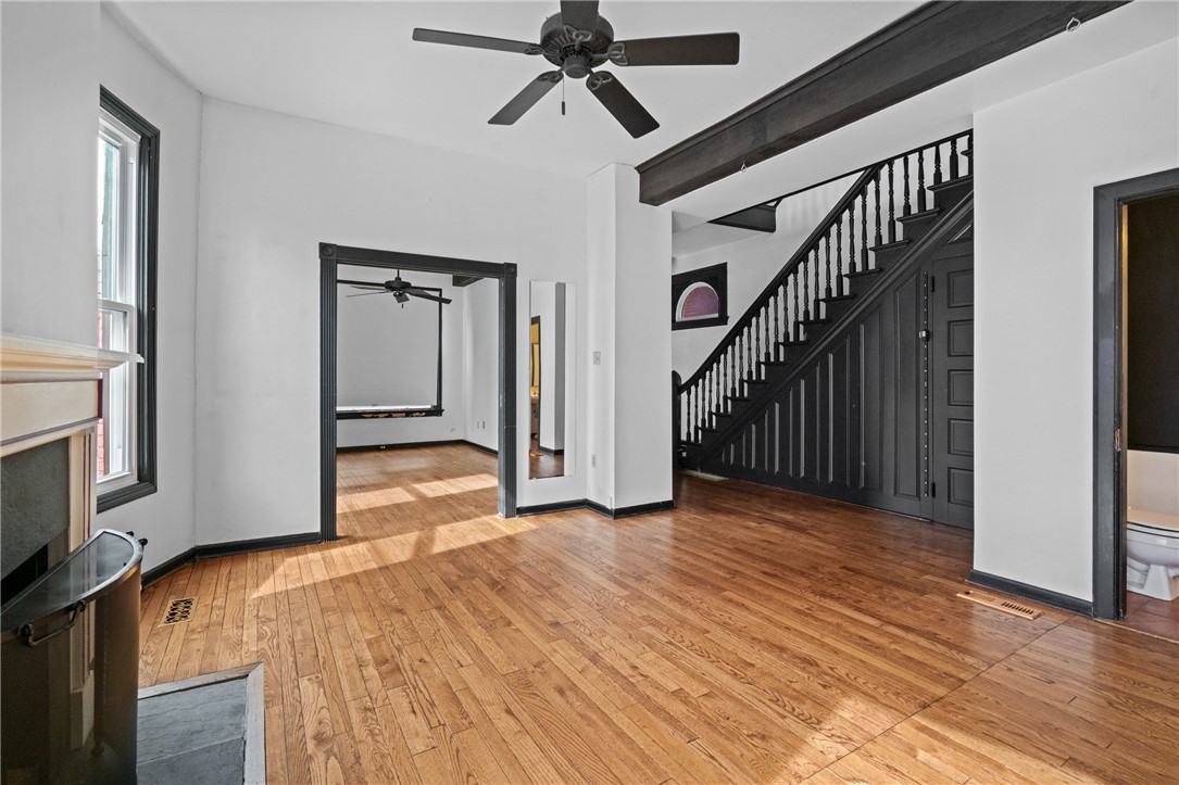 Empty room with hardwood floors, a dark staircase, exposed beam, and ceiling fan.