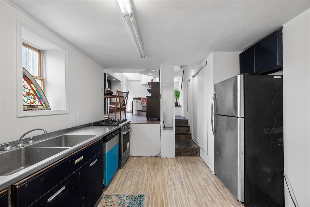 Narrow galley kitchen with a stained-glass window, stainless-steel sink and refrigerator, and steps to a dining area.