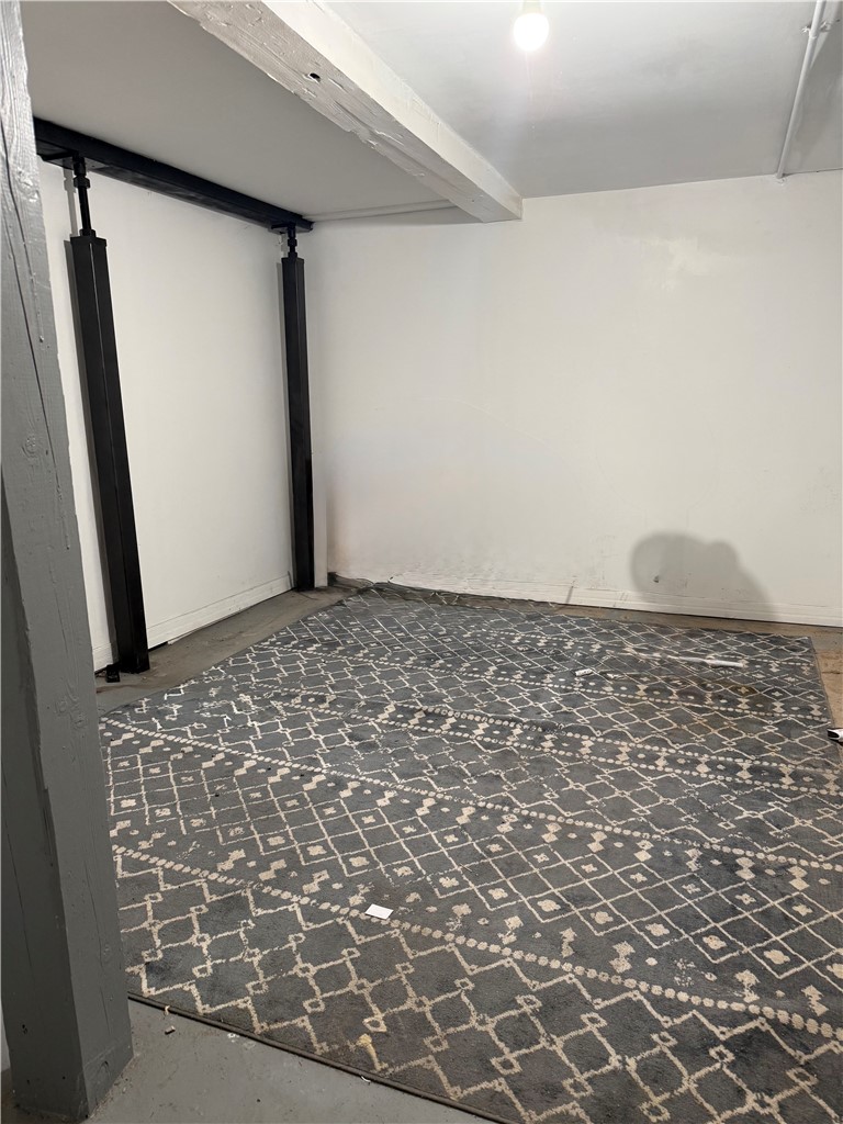 Basement room with exposed beams, black support posts, and a gray patterned rug.