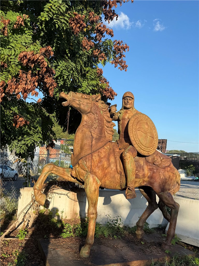 Weathered statue of an armored rider on a rearing horse beneath a leafy tree.