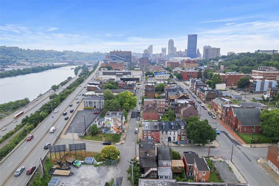Aerial view of an urban neighborhood with the Pittsburgh skyline, river, and highway beyond.