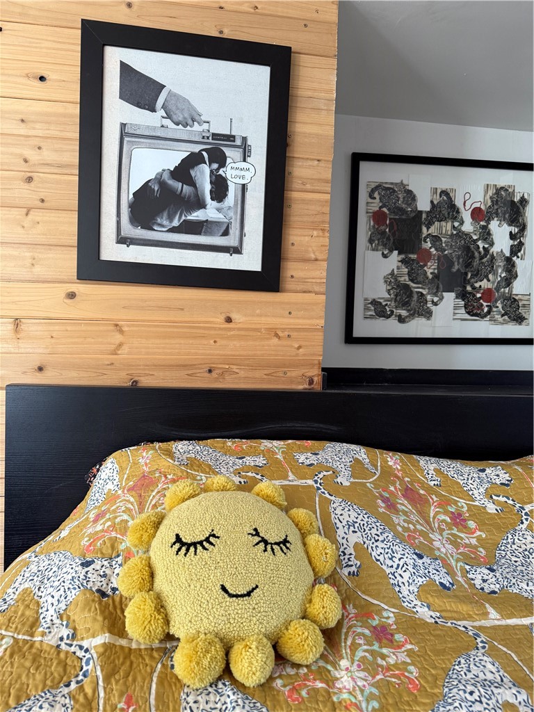 Bedroom with a patterned quilt, yellow smiling sun pillow, wood-plank wall, and framed artwork.