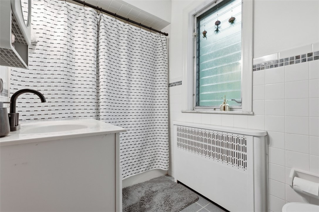 Tiled bathroom with a white vanity, patterned shower curtain, frosted window, and radiator cover.