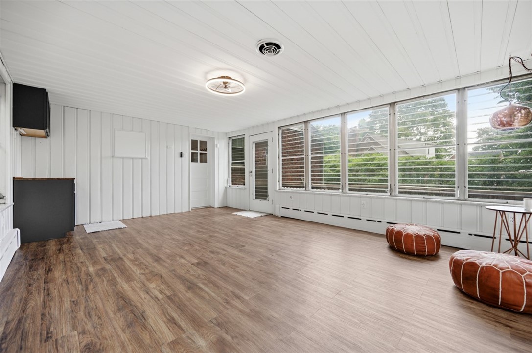 Enclosed sunroom with wide windows, white paneling, wood-look floors, and two rust-colored poufs.