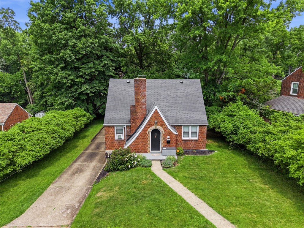 Aerial view of a red-brick house with a steep roof and chimney, surrounded by mature trees and a green lawn.