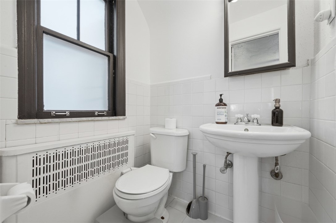 White-tiled bathroom with a pedestal sink, toilet, and dark-trimmed window.