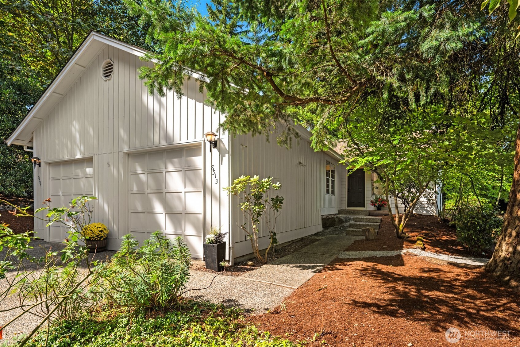 White-sided home with two garage doors, a side entrance, and a tree-shaded garden walkway.