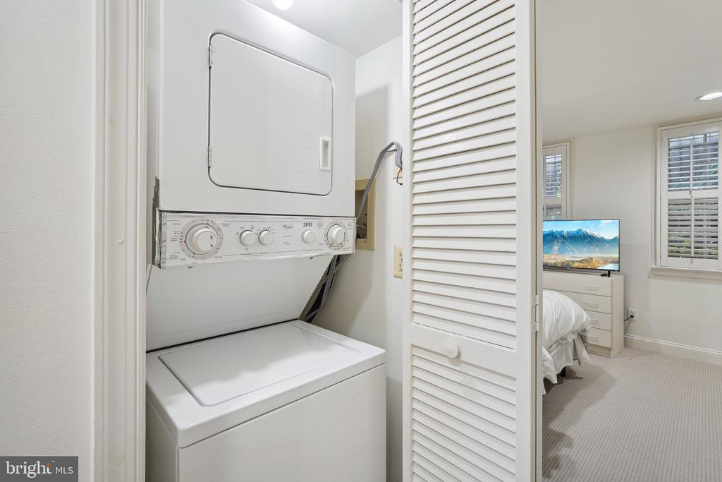Stacked washer and dryer in a louvered closet beside a bright bedroom.