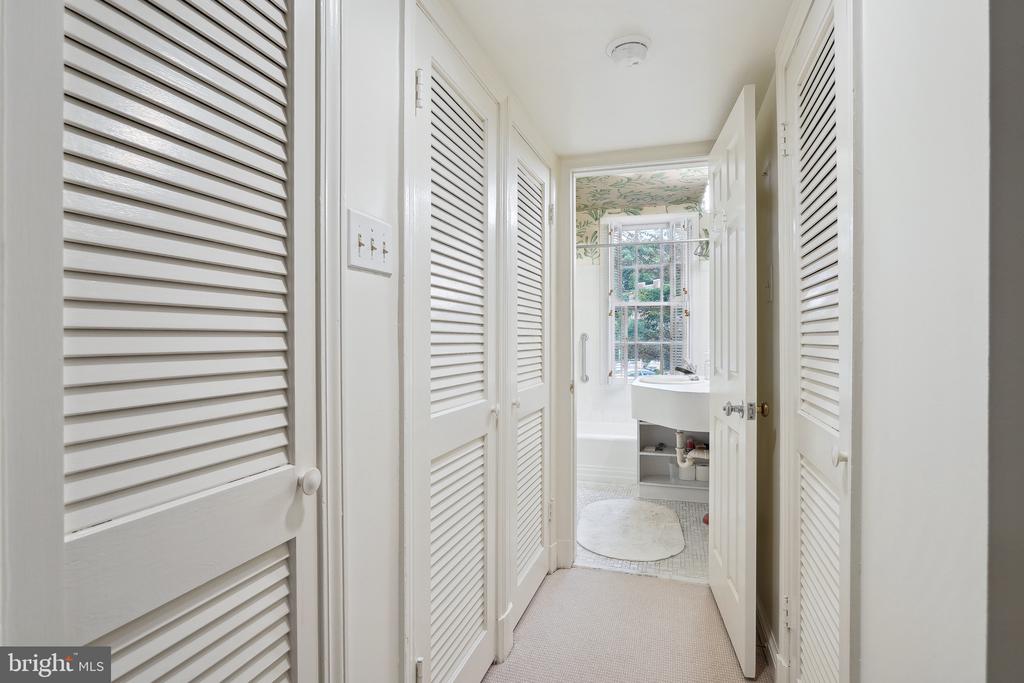 Hallway with white louvered closet doors leading to a bathroom with a pedestal sink and window.